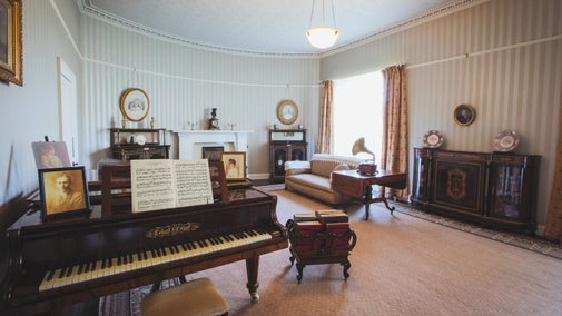 A front room with a piano, fireplace, gramophone and long pink curtains against stripey wallpaper
